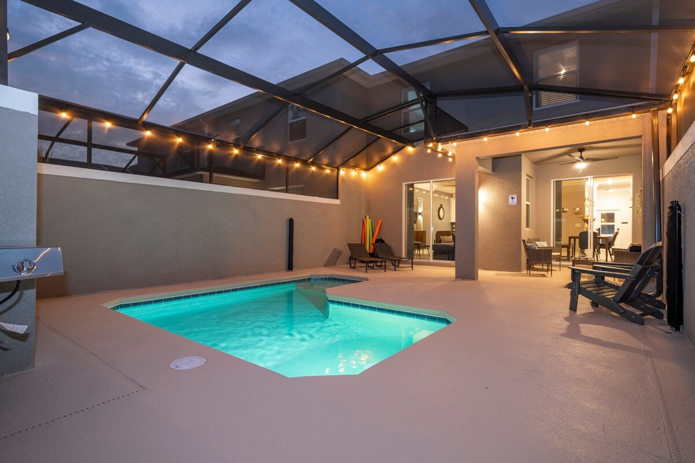 A spacious, comfortable, and family-friendly outdoor pool area that is perfect for any guest seeking the ultimate vacation experience. Imagine yourself unwinding in a large, inviting pool area, surrounded by soft lighting that creates an enchanting evening atmosphere. The space is designed with modern aesthetics and convenience in mind, ensuring every moment of your stay is steeped in comfort and luxury. The pool area is enclosed by a screened structure, and there are several chairs arranged around the pool for relaxation or socializing. A grill is mounted on one of the walls, indicating an outdoor cooking area. The floor is uniformly covered with light-colored tiles giving it a clean and elegant look. Large glass doors lead into the interior of a house, suggesting that this pool area is part of a residential property.